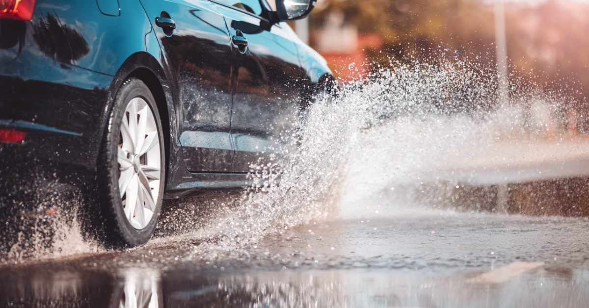 A car tire splashing through a large puddle on a wet road, with water spraying around the wheel in rainy weather.