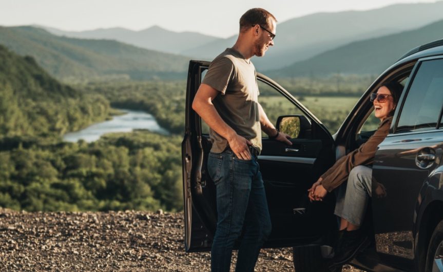 A person standing next to a car and talking to a person sitting in the driver’s seat. The car is parked by a forest.