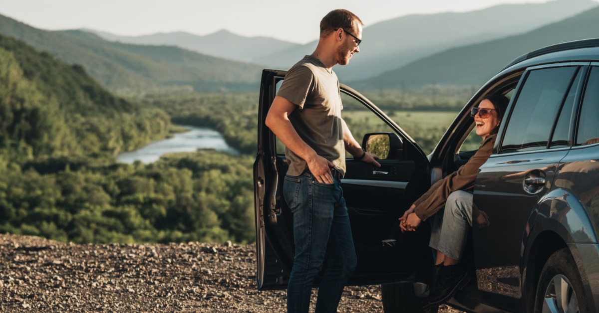 A person standing next to a car and talking to a person sitting in the driver’s seat. The car is parked by a forest.