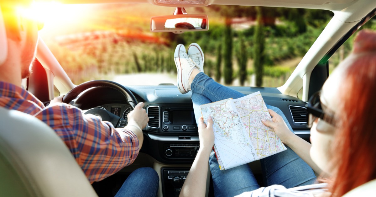 Two people in a car driving near a large forest. The passenger has their shoes on the dashboard and holds a map.