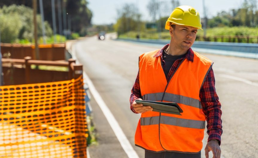A construction worker holding a tablet and looking down the road. The worker stands next to a construction zone outside.