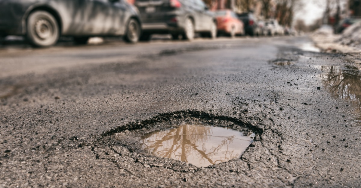 Many cars parallel parked on a long street outside. A large pothole full of muddy water sits in the middle of the road.