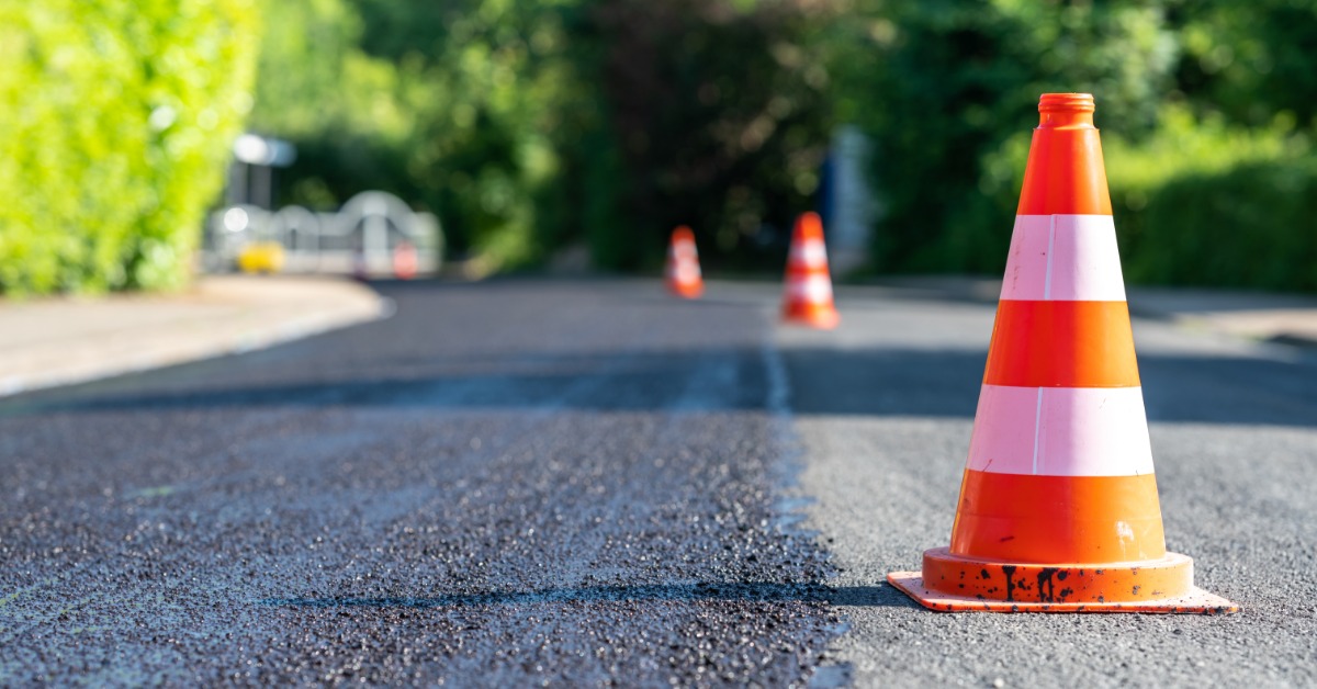 Three orange construction cones sitting in a road under construction. Half the road is new asphalt, and the other half is still old material.