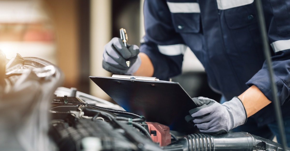 A close-up of a man with a clipboard in a work suit and gloves writing while leaning over the open engine bay of a car.
