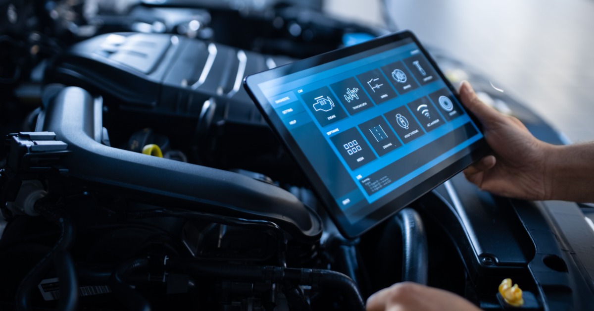 A close-up of a person holding an electronic tablet with automotive icons over the open engine bay of a car.
