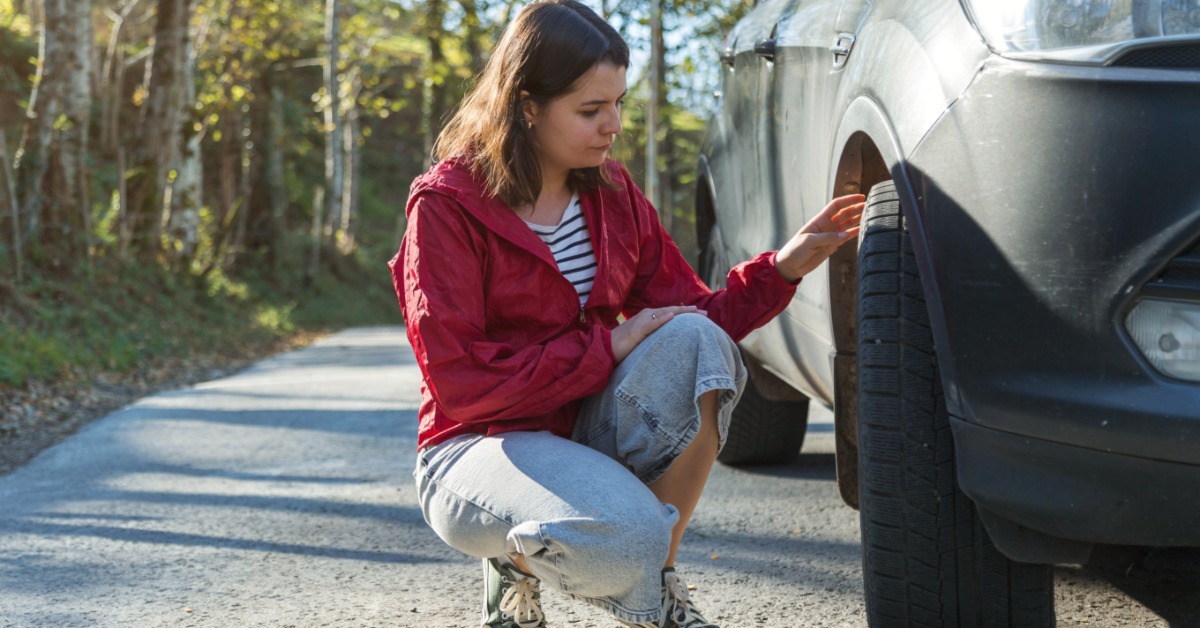 A person wearing a burgundy jacket leans near the tire of a car on the side of a road. Sunlight shines on the gray car.