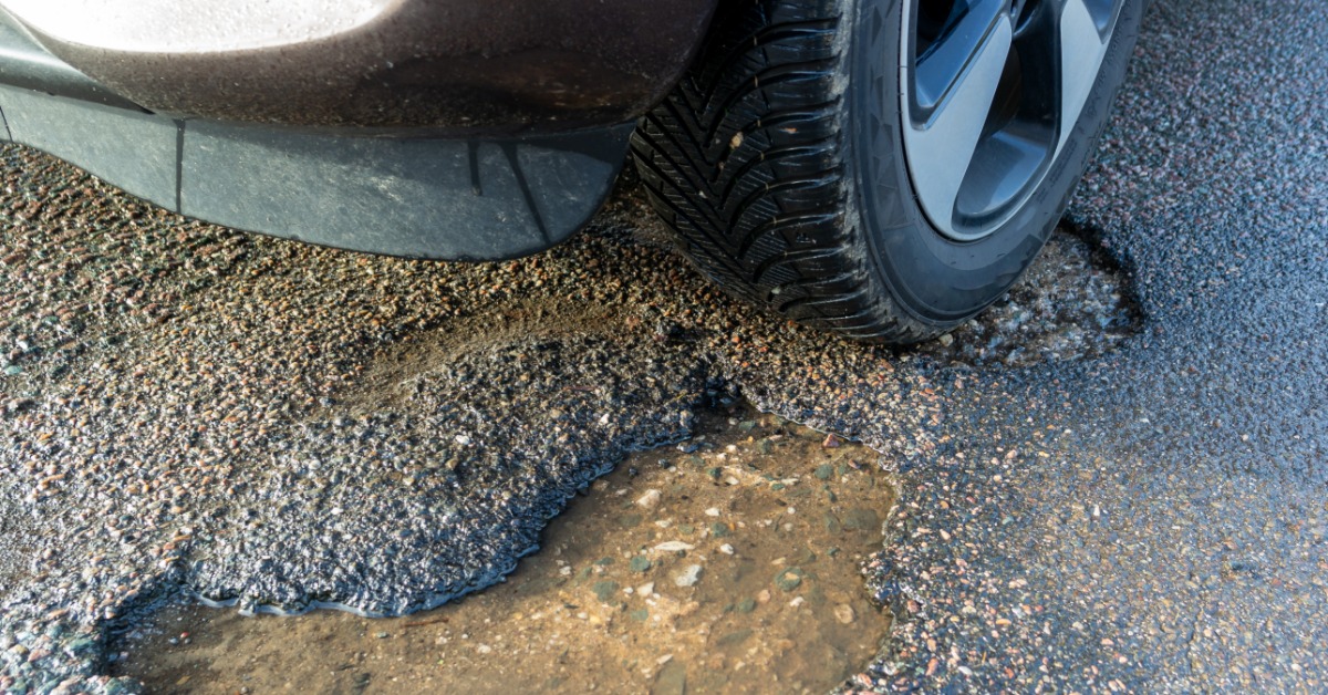 A pothole in cracked asphalt beside a car tire on a road surface with loose stones and rough pavement.
