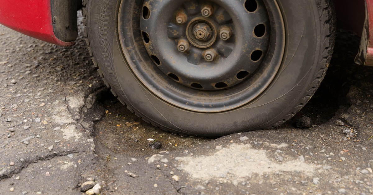 The flat tire of a red car is resting on damaged asphalt with cracks, rough pavement, and road debris nearby.