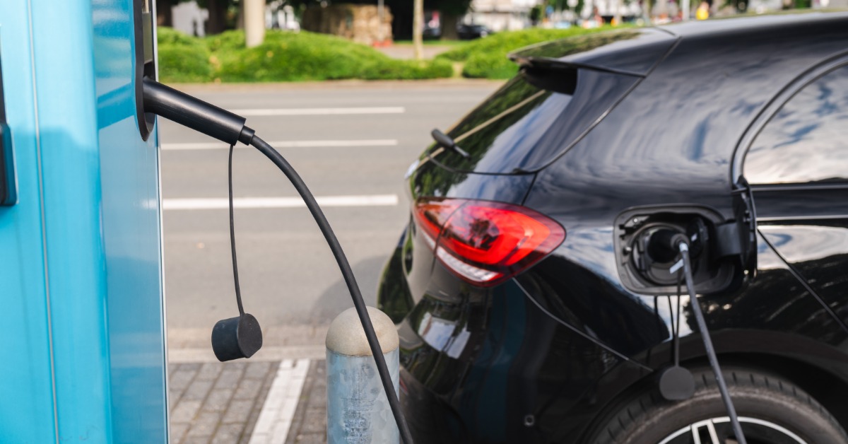 A black electric car connected to a charging cable at the rear port, with the rear light and tire visible.