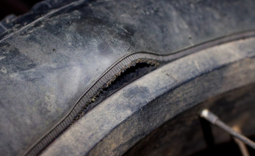 A ruptured tire showing torn sidewall rubber, exposed cord fabric, thick tread, and dark debris nearby.