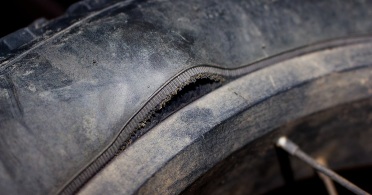 A ruptured tire showing torn sidewall rubber, exposed cord fabric, thick tread, and dark debris nearby.