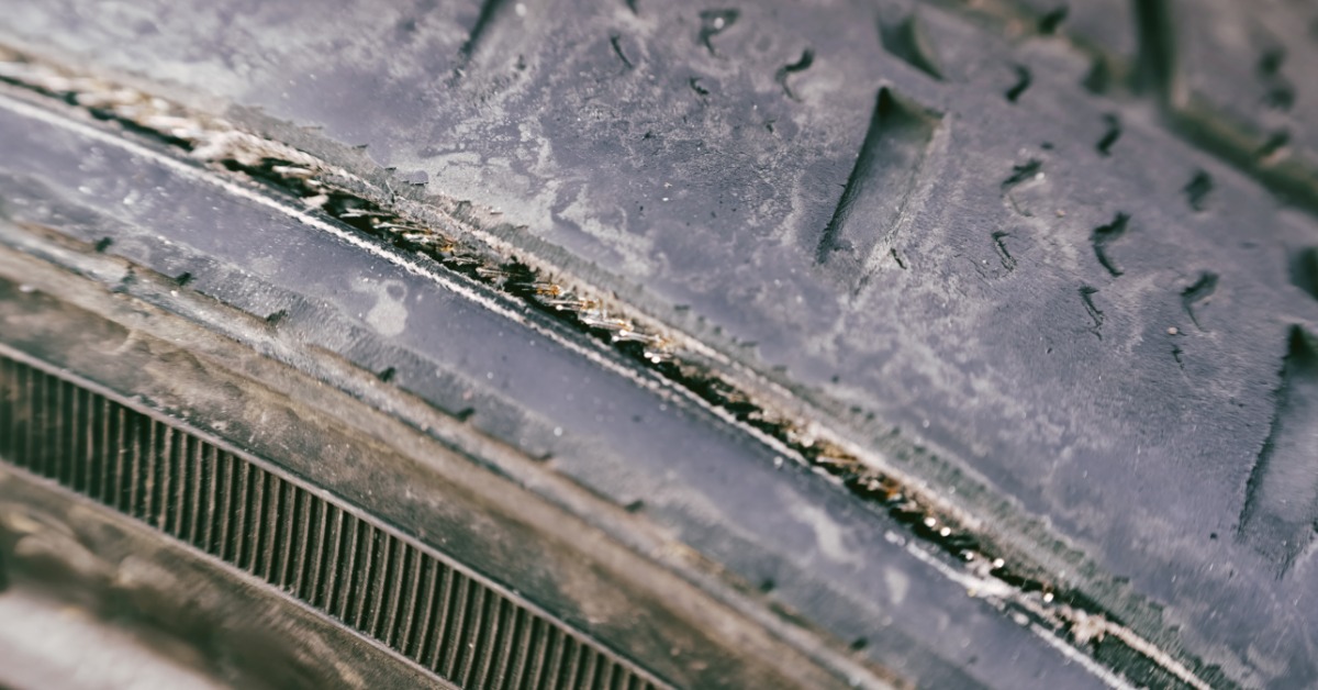 A used vehicle tire with damaged rubber, worn tread grooves, rough surface texture, and dark background blur.