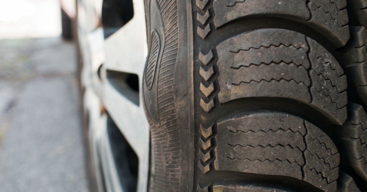 A car tire sidewall with a raised bulge, black rubber texture, tread edges, and a blurred pavement background.
