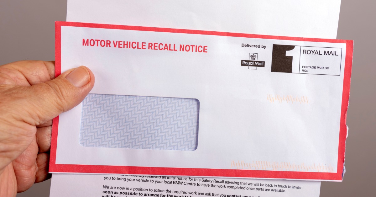 A close-up of a hand holding a mail envelope with a red border that says "MOTOR VEHICLE RECALL NOTICE."