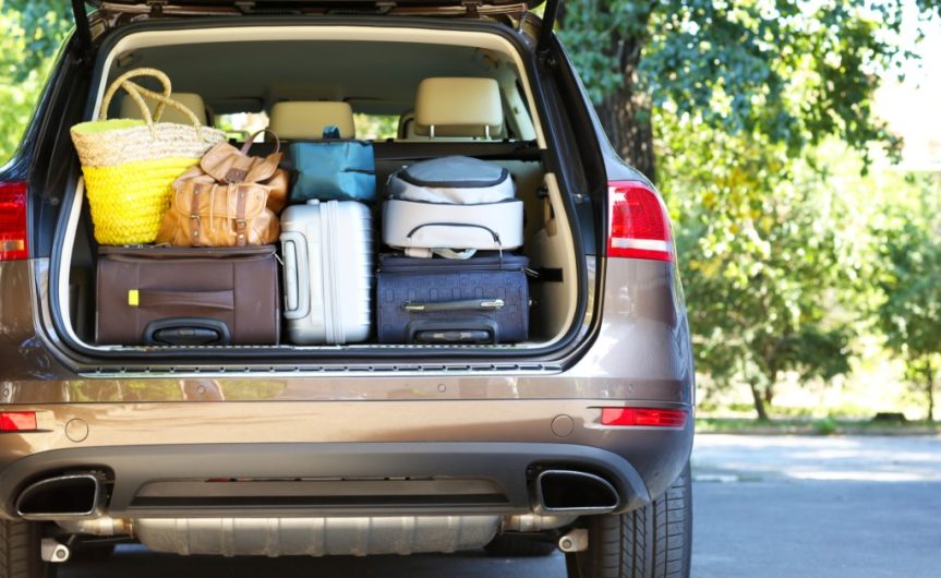 A large beige car with its back door fully open on a sunny day. The car trunk is full of stacked luggage.