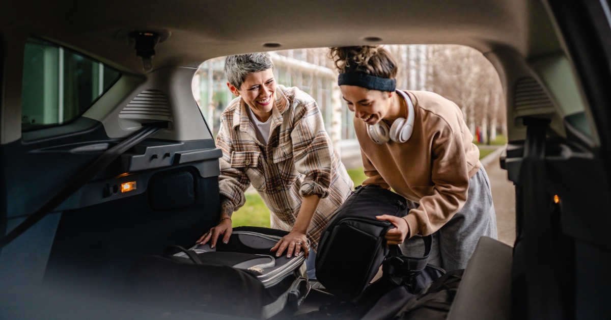 Two people loading luggage into the large trunk of a car during the daytime. Both people laugh while packing.
