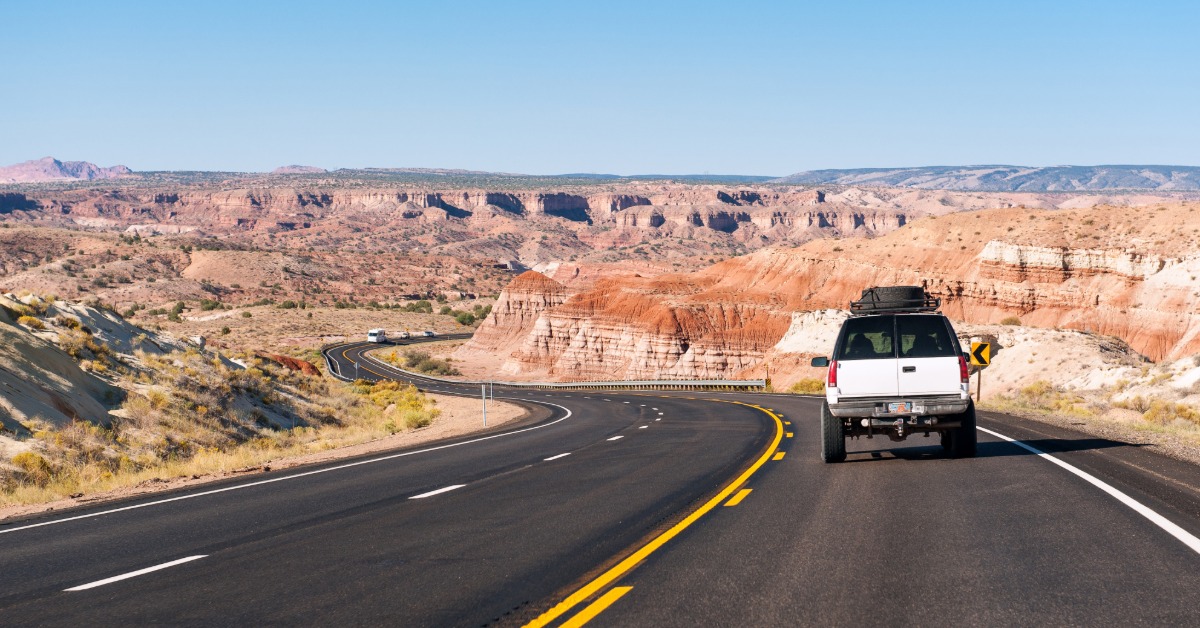 A large white car driving on a long stretch of empty road in Arizona on a sunny day. The car has roof cargo.