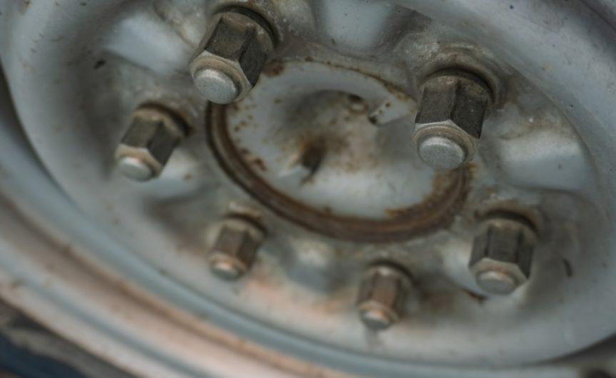 A tire attached to a dirty, white car wheel with six large, metal lug nuts. The wheel is partially rusted.