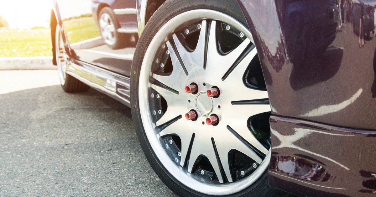 A burgundy car parked outside on the street on a bright, sunny day. The front driver-side wheel has four red lug nuts.