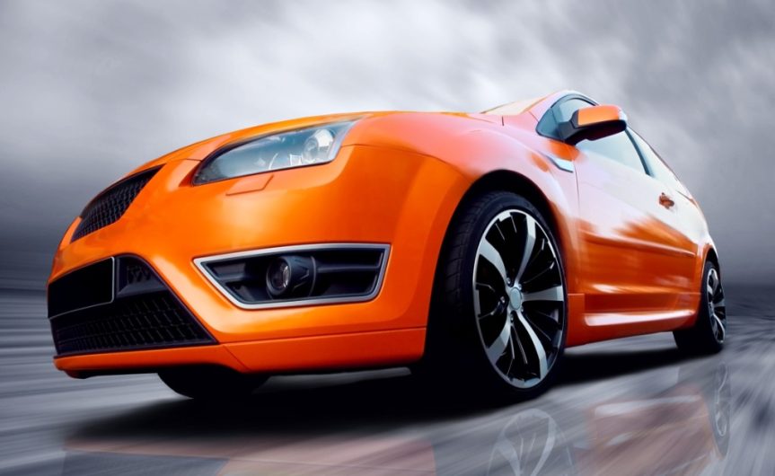 A low-angle, frontal view of an orange two-door sports car driving on a grey surface underneath a grey sky.