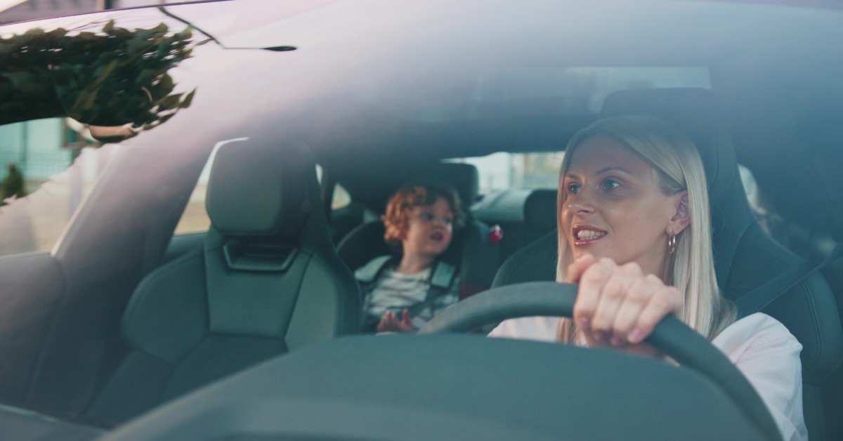 A blonde adult woman is driving a car and looking in the rearview mirror to talk to a toddler boy in the back seat.