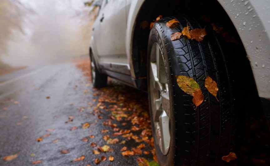 A large gray car driving on wet road on a foggy day. The road is covered in orange and brown fallen leaves.