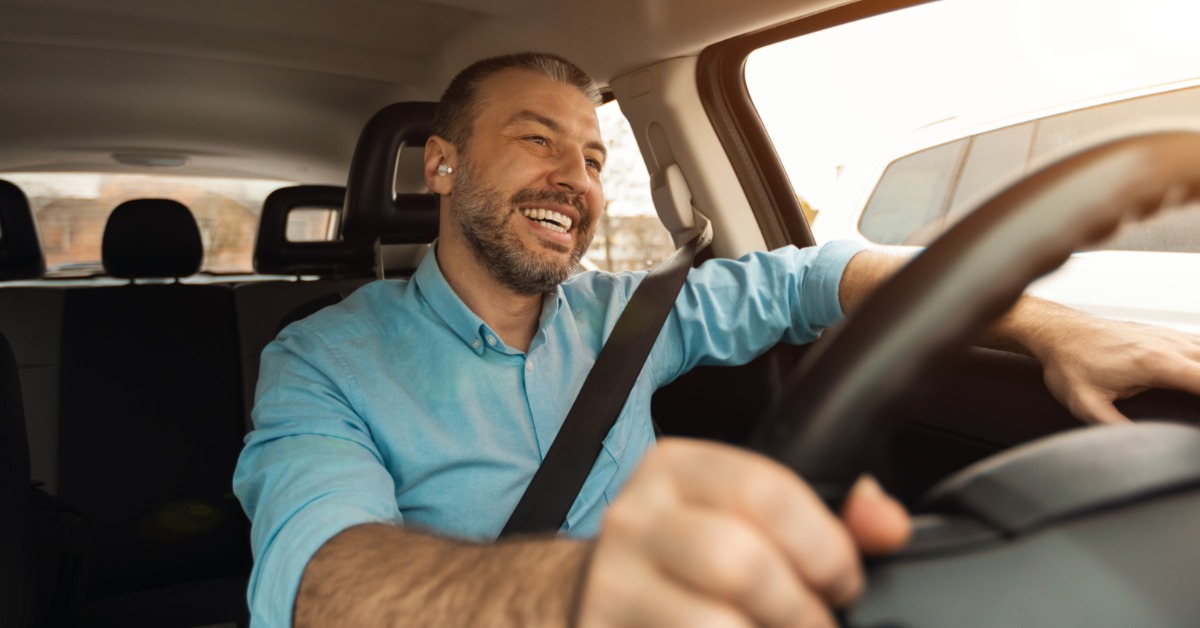A person wearing a light blue shirt and white air pods smiles while driving a car during the daytime.