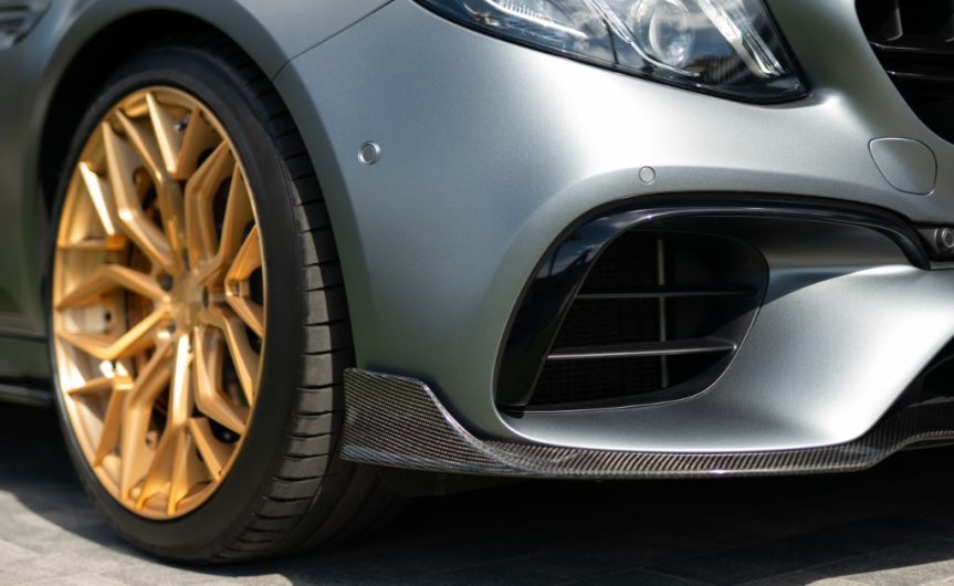 A close-up of the front bumper and wheel of a matte grey sports car with gold alloy wheels outside on a sunny day.