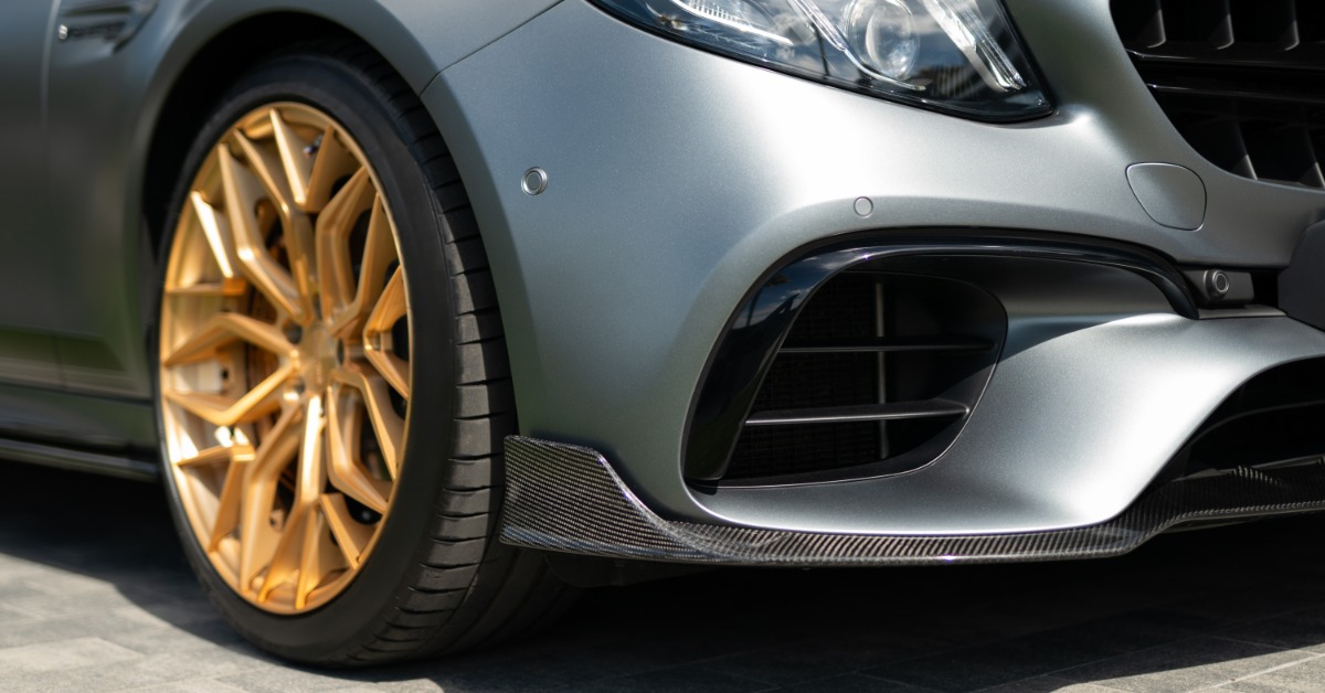 A close-up of the front bumper and wheel of a matte grey sports car with gold alloy wheels outside on a sunny day.