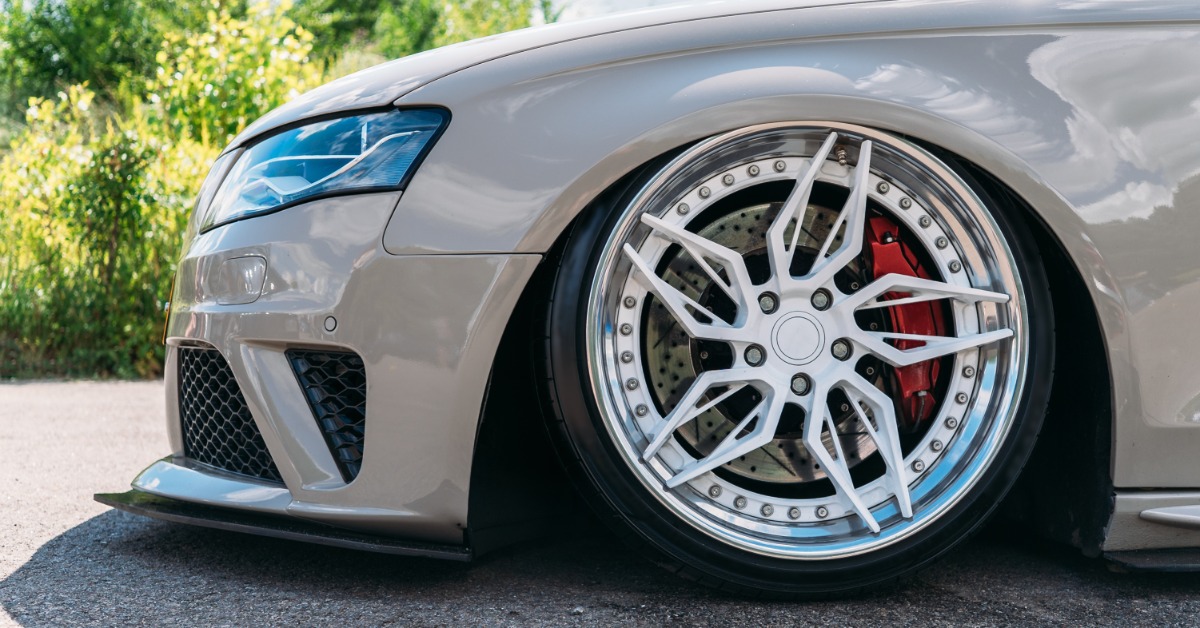 A close-up of the front custom wheel and tire of a beige lowrider sports car with red brake calipers.