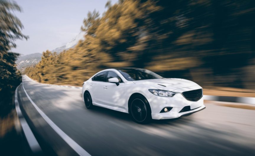 A front and rear view of a white sedan with black wheels driving on a single-lane asphalt road during the daytime.