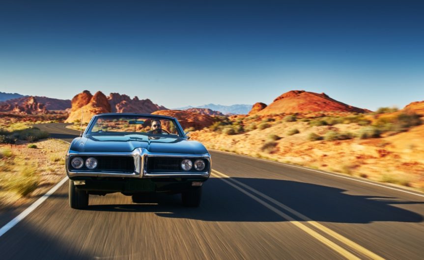 A person driving a vintage car with the top down on a road running through a large desert during the daytime.