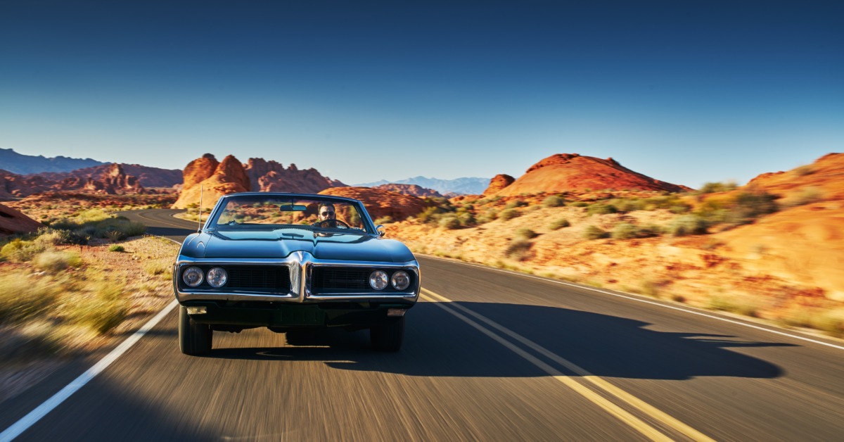 A person driving a vintage car with the top down on a road running through a large desert during the daytime.
