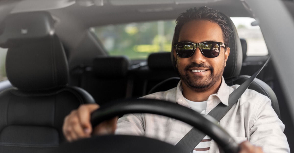 A person wearing sleek, black sunglasses and a striped shirt smiles while driving a car near trees outside.