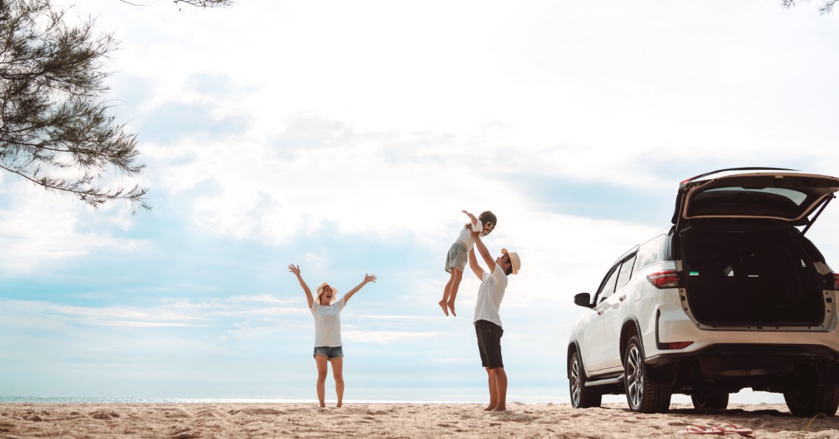 A parent holding their child up in the air next to their car on a beach. The other parent stands nearby.