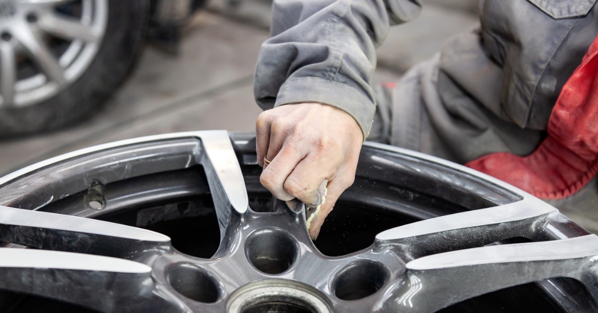 A close-up of a man in a work suit cleaning the surface of a detached automotive aluminum wheel rim with a cloth.