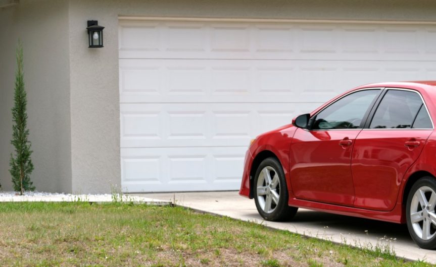 A small red car parked in the driveway of a beige house with a wide, white garage during the daytime.