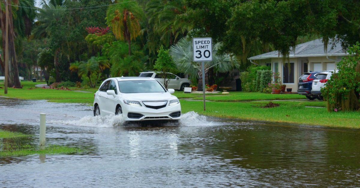 A white car driving through a flooded street on a gray, cloudy day. The street is surrounded by tall trees.