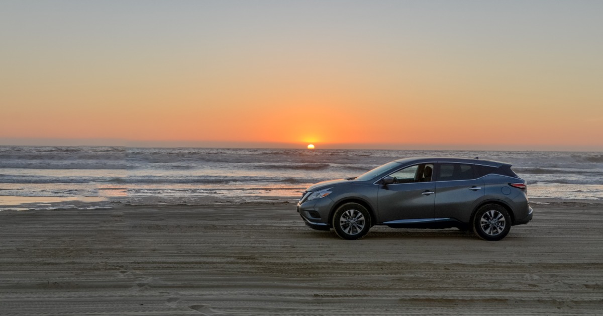 A large gray car driving on the beach during sundown. The orange sun is barely visible across the ocean.