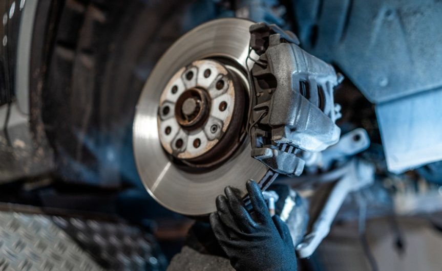 A close-up of a vehicle's exposed brake disc and caliper as a hand with a black glove touches it in a workshop.