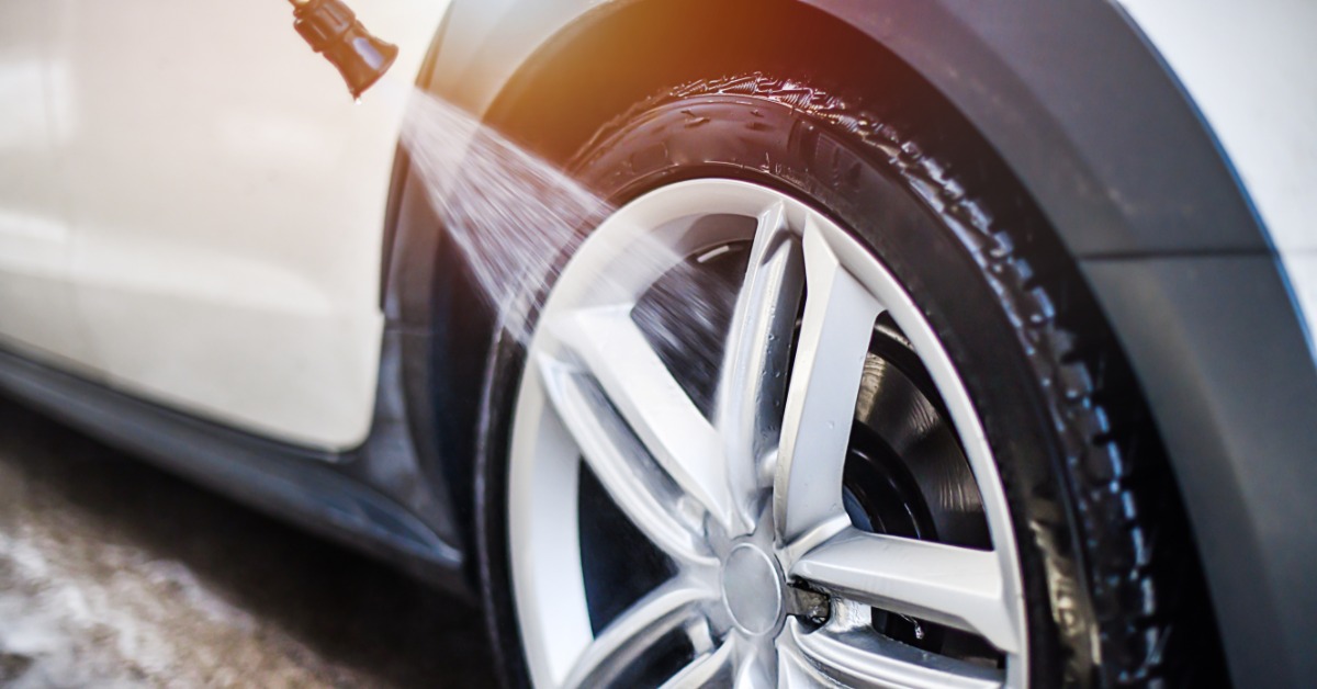 A close-up of the wheel and tire of a white car being sprayed by a water jet outside on a sunny day.