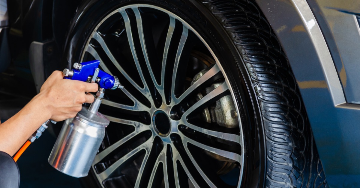 A close-up of a hand using a stainless-steel spray bottle attached to a hose to apply a spray coating to a car wheel.
