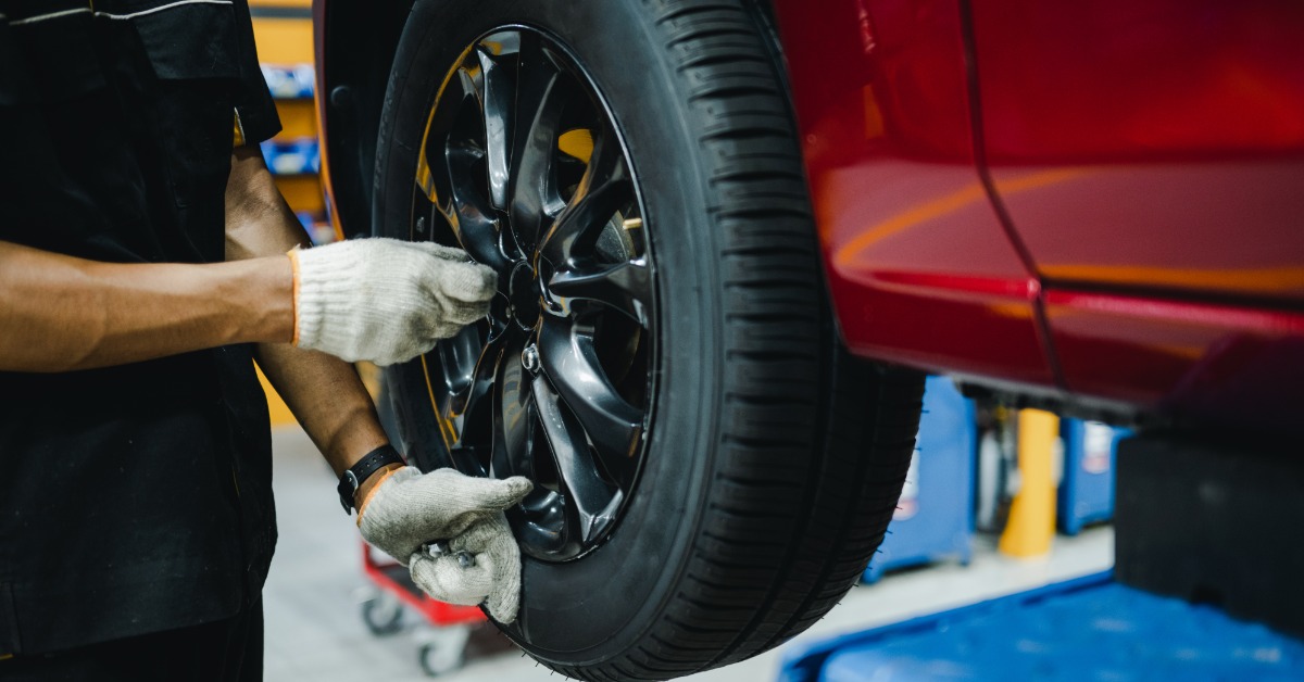 A close-up of a man's hands with white gloves inspecting the wheel of a red car elevated on a lift in a shop.