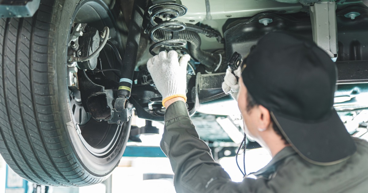 A rear view of a man with a black baseball hat and white gloves inspecting the springs of a car's suspension.