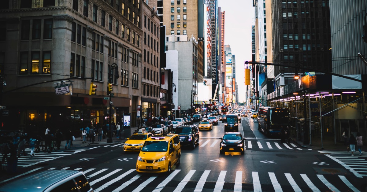 A city street full of cars driving next to tall skyscrapers. Many people cross the street on a nearby crosswalk.