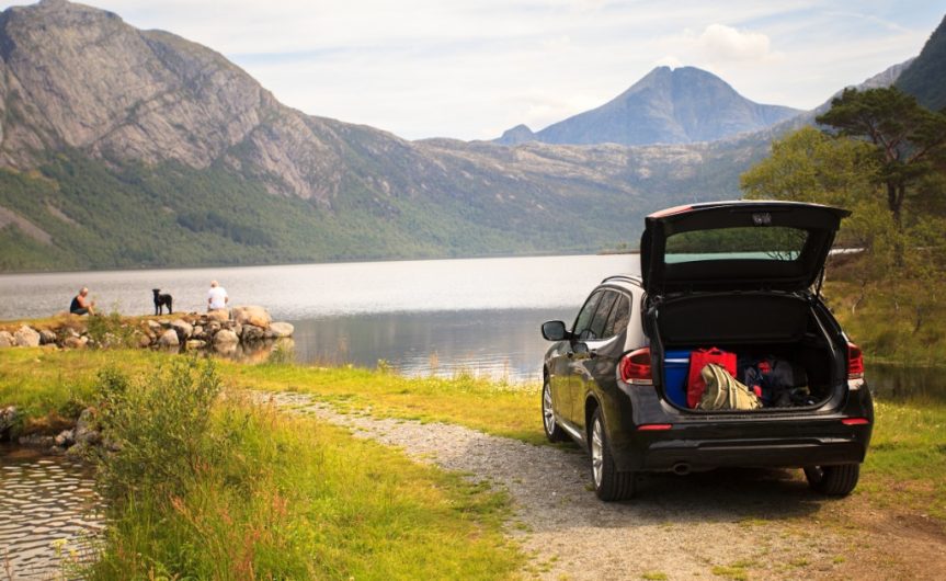 A rear view of a car parked next to a lake with the trunk open and luggage inside while two people and a dog sit nearby.