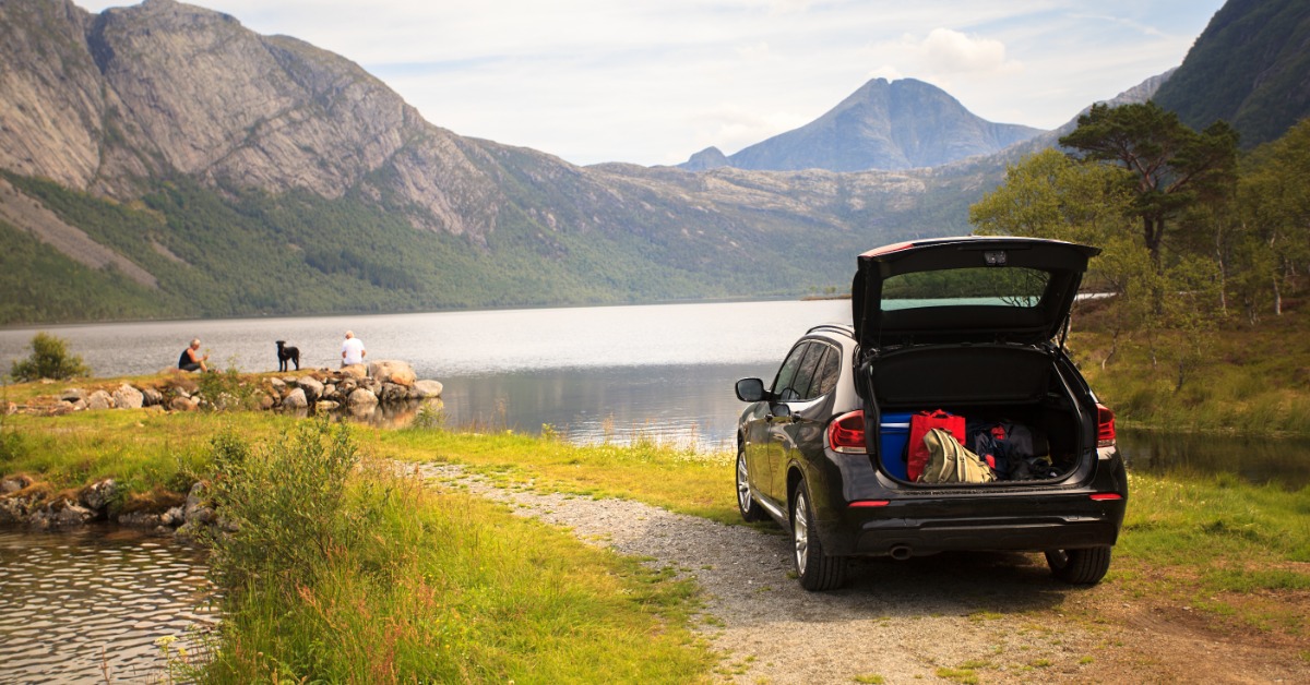 A rear view of a car parked next to a lake with the trunk open and luggage inside while two people and a dog sit nearby.