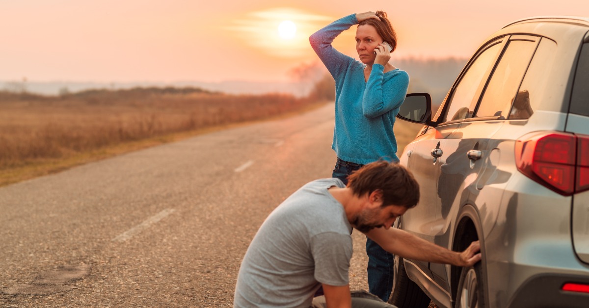 A woman standing and talking on the phone next to a car parked on the side of the road while a man crouches near the tire.