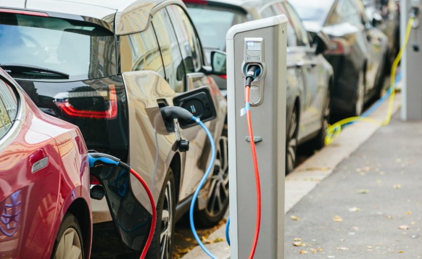 A line of different electric vehicles parked outside during the daytime. The cars are attached to EV chargers.