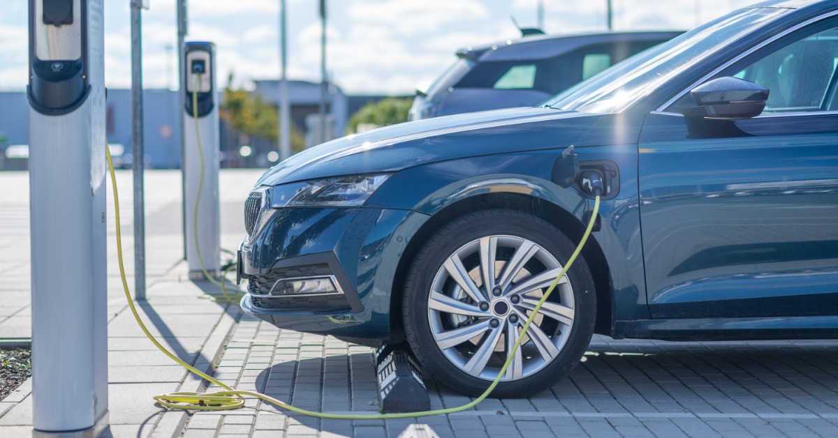 Two dark gray electric vehicles in a parking lot on a sunny day. Both cars are hooked up to EV chargers on a sidewalk.